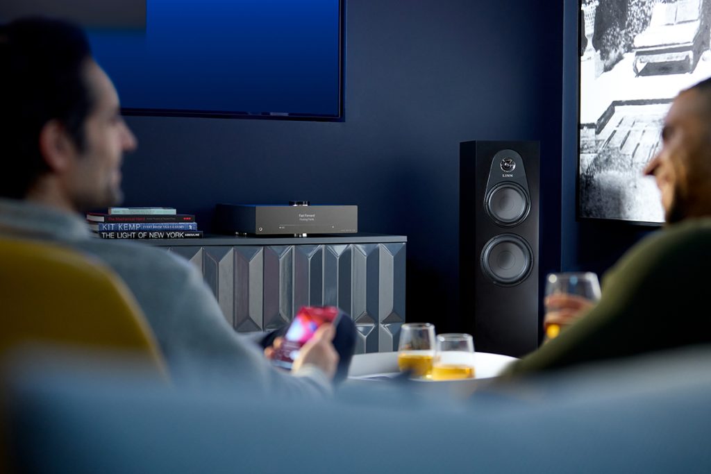 Background: Linn Majik DSM/5 next to a Linn 150 loudspeaker. Foreground: two people sitting. One holding a phone with the Linn App open.
